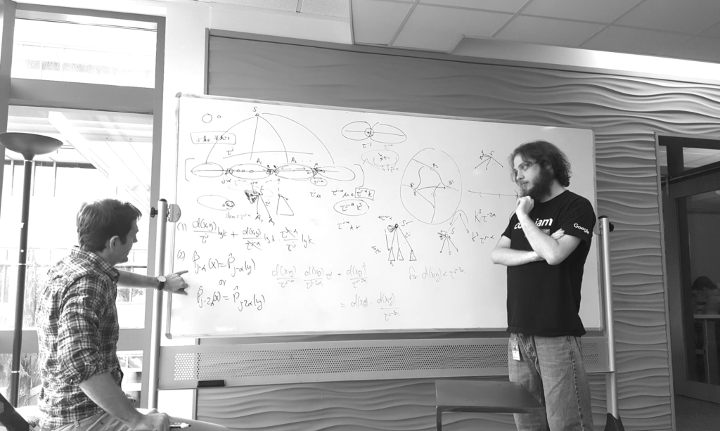 Foto en blanco y negro que muestra a Mikey trabajando con un colega cerca de una pizarra blanca cubierta de ecuaciones.